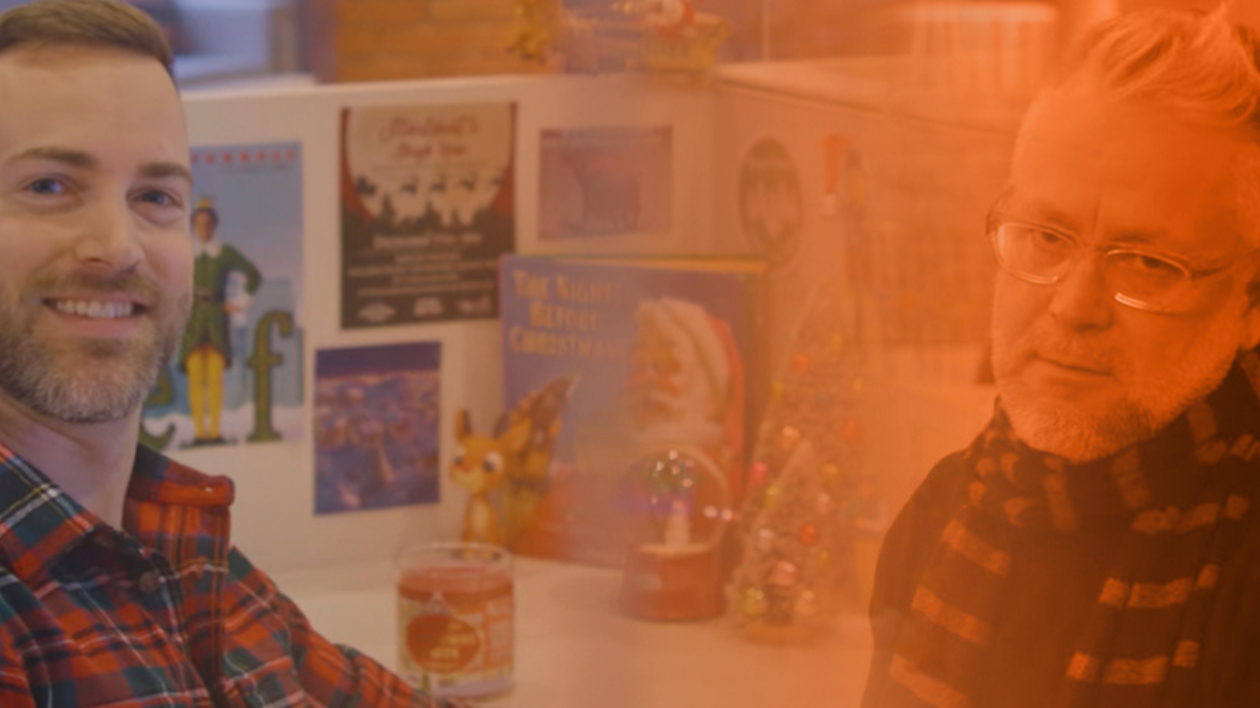 Image of two people sitting in a cubile one with an annoyed expression. The other with a smile.