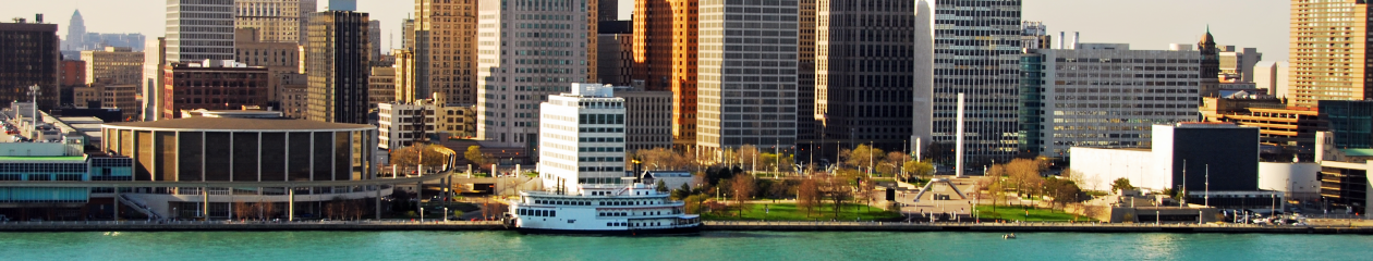 Boats in the Detroit River are seen in the foreground with the Detroit riverfront and skyline in the background, on a sunny summer day.