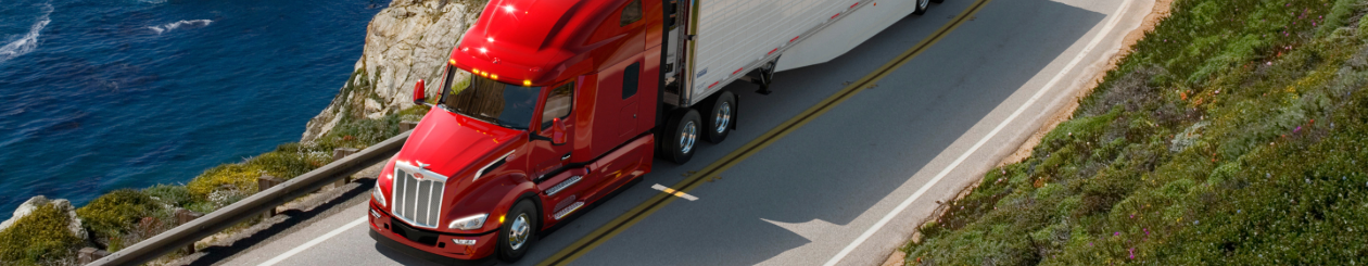 A shiny Peterbilt semi truck travels along an ocean-view highway with greenery all around