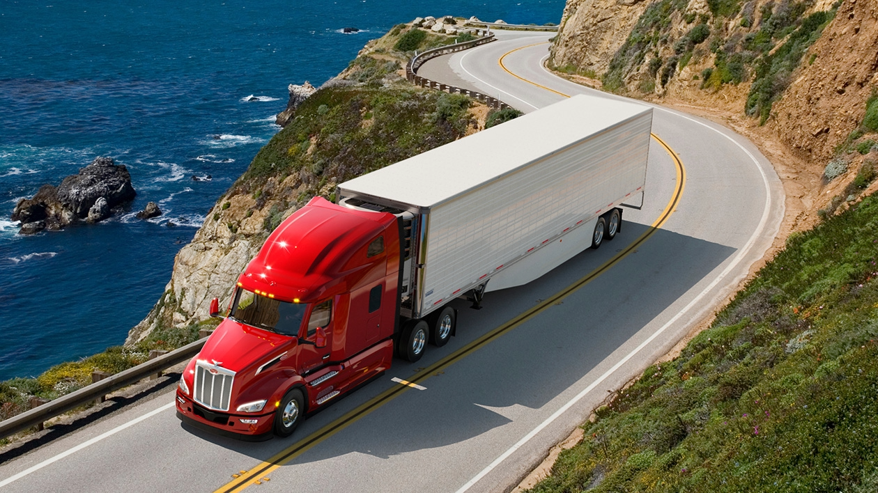 A shiny Peterbilt semi truck travels along an ocean-view highway with greenery all around