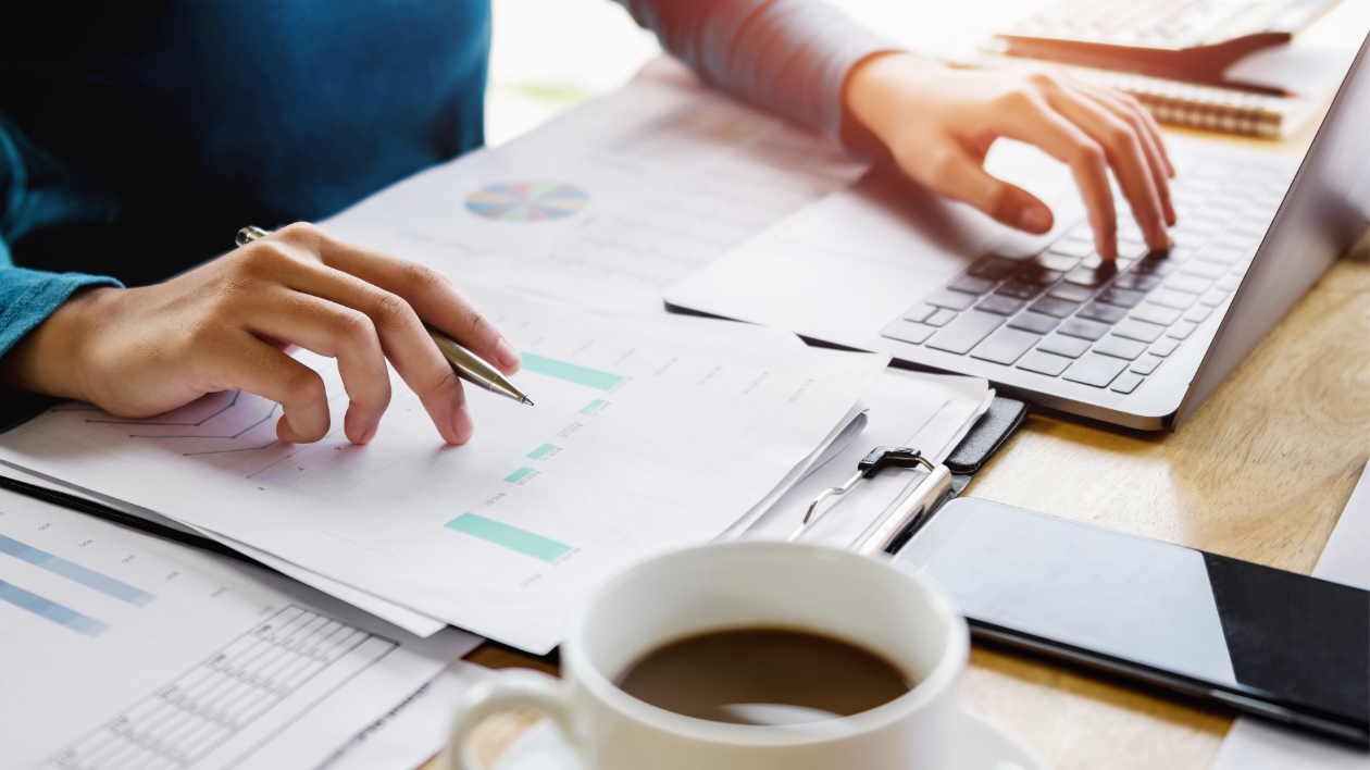 An individual sits at a wooden desk or table with a cup of coffee, paperwork, a mobile device and computer as they are busy at work, writing on a notepad